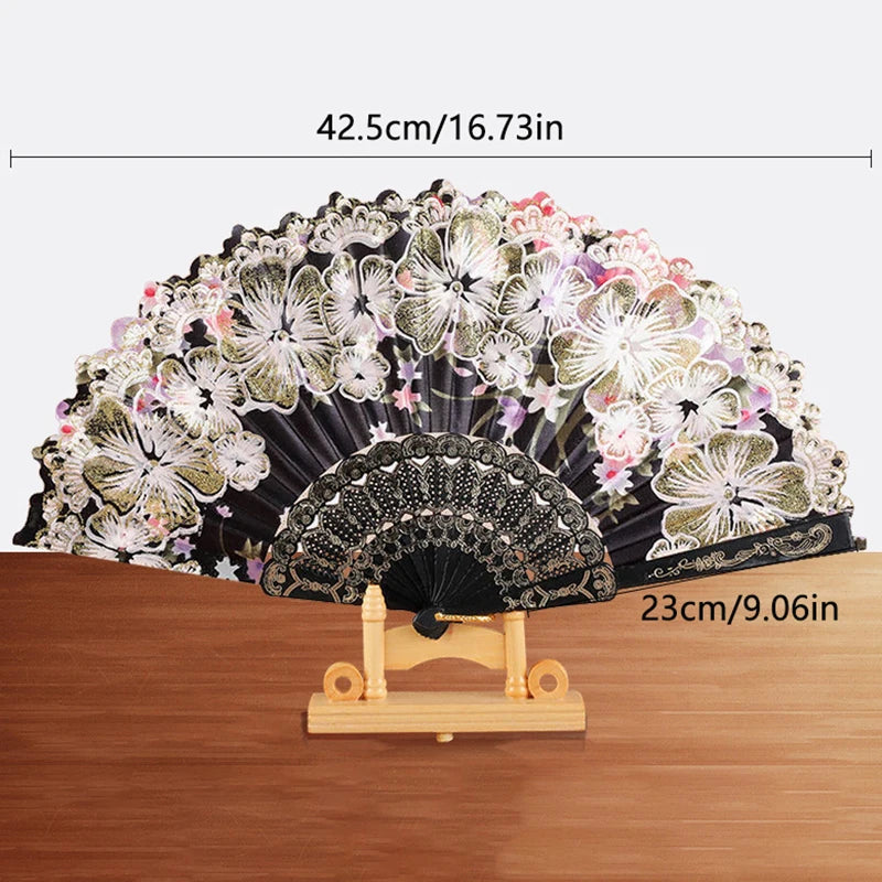 A black silk flower folding fan with pink and white floral patterns displayed on a wooden surface.
