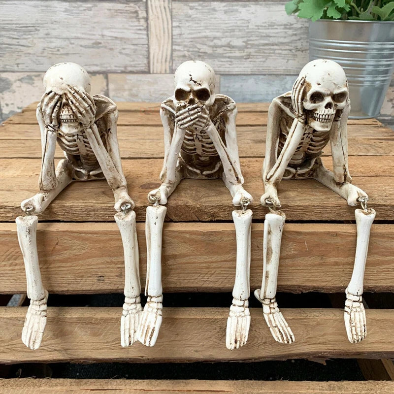 A set of three white resin skeleton skull figurines posed with hands to their face on a wooden surface.