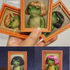 A hand holds an open wooden photo frame displaying a painting of a green frog with blonde hair.