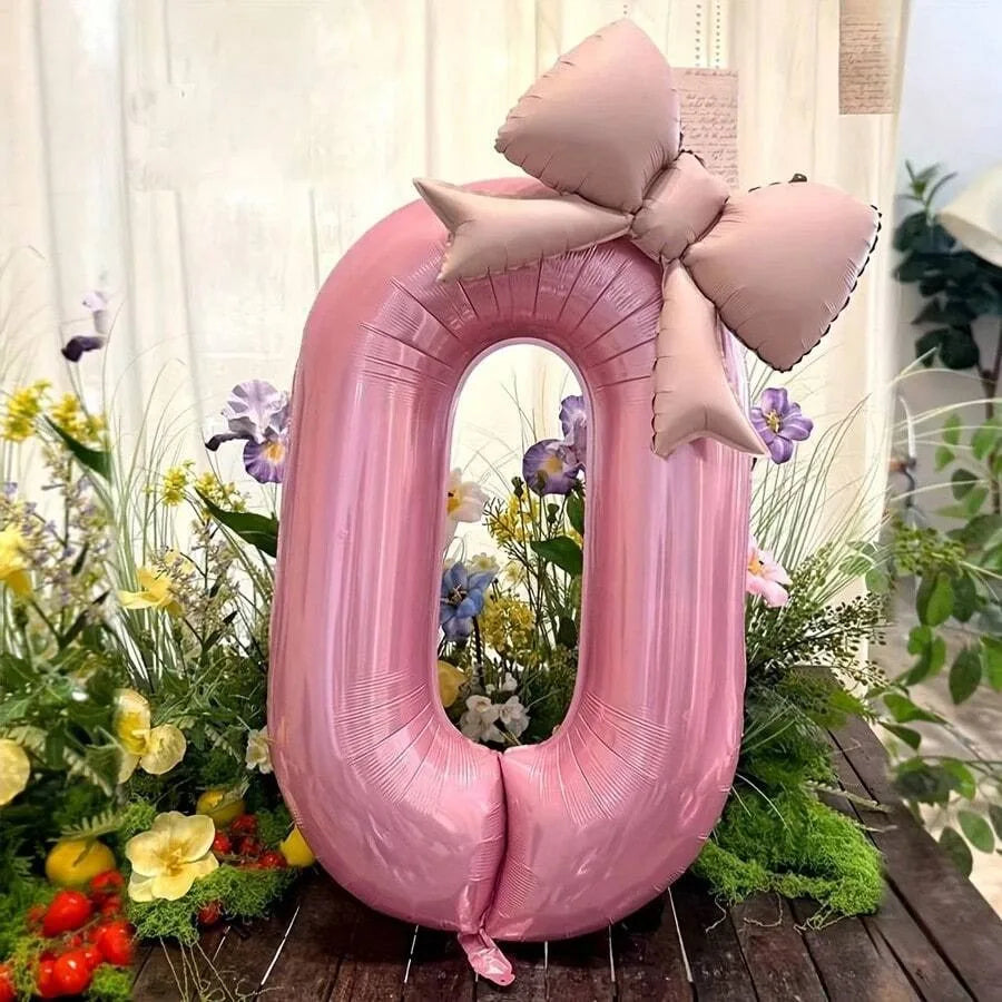 A large pink foil balloon shaped like the number zero decorated with a matte pink bow surrounded by greenery and flowers.