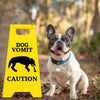 A light-colored dog sitting next to a yellow caution sign that reads DOG VOMIT CAUTION
