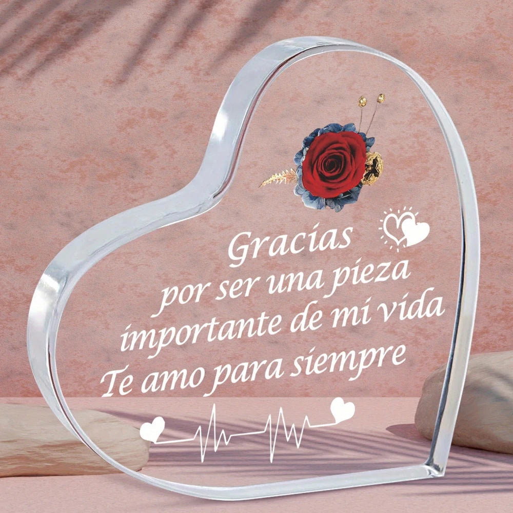 A clear glass heart-shaped keepsake frame engraved with Spanish text and a red rose flower.