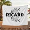 A white flag featuring the name RICARD in black text with a sunburst design, displayed outdoors.