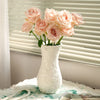 A white ceramic vase filled with pink roses sits on a white cloth next to a window blind.
