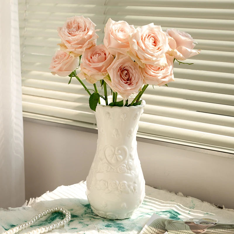 A white ceramic vase filled with pink roses sits on a white cloth next to a window blind.