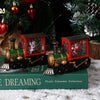 A miniature Christmas train music box with red and green accents sits on a green book in a snowy setting.