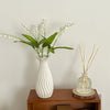 A white ceramic bud vase holds delicate white flowers and green leaves on a wooden surface.