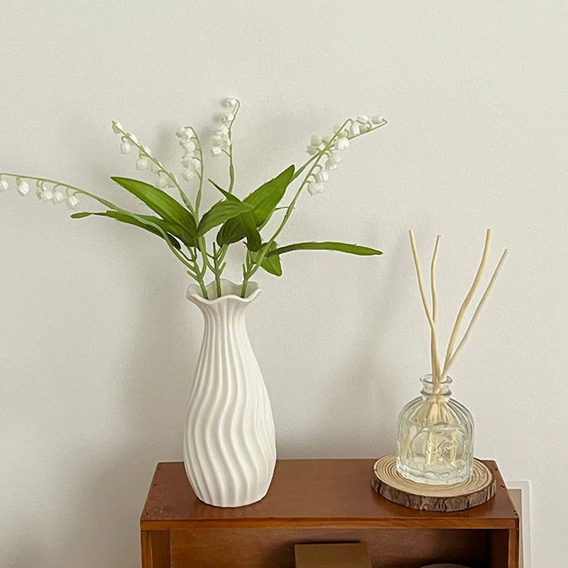 A white ceramic bud vase holds delicate white flowers and green leaves on a wooden surface.