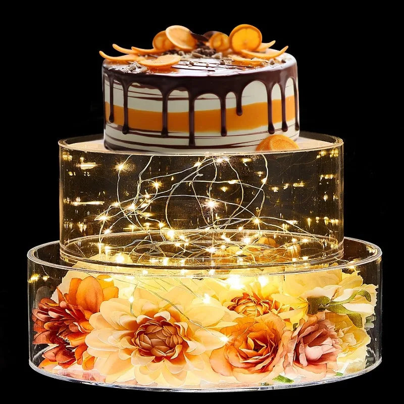 A tiered cake display featuring an orange and white cake under a clear acrylic stand illuminated by warm LED lights.