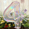 A large, iridescent, transparent foil balloon shaped like the number 4 is decorated with a bow and surrounded by greenery.