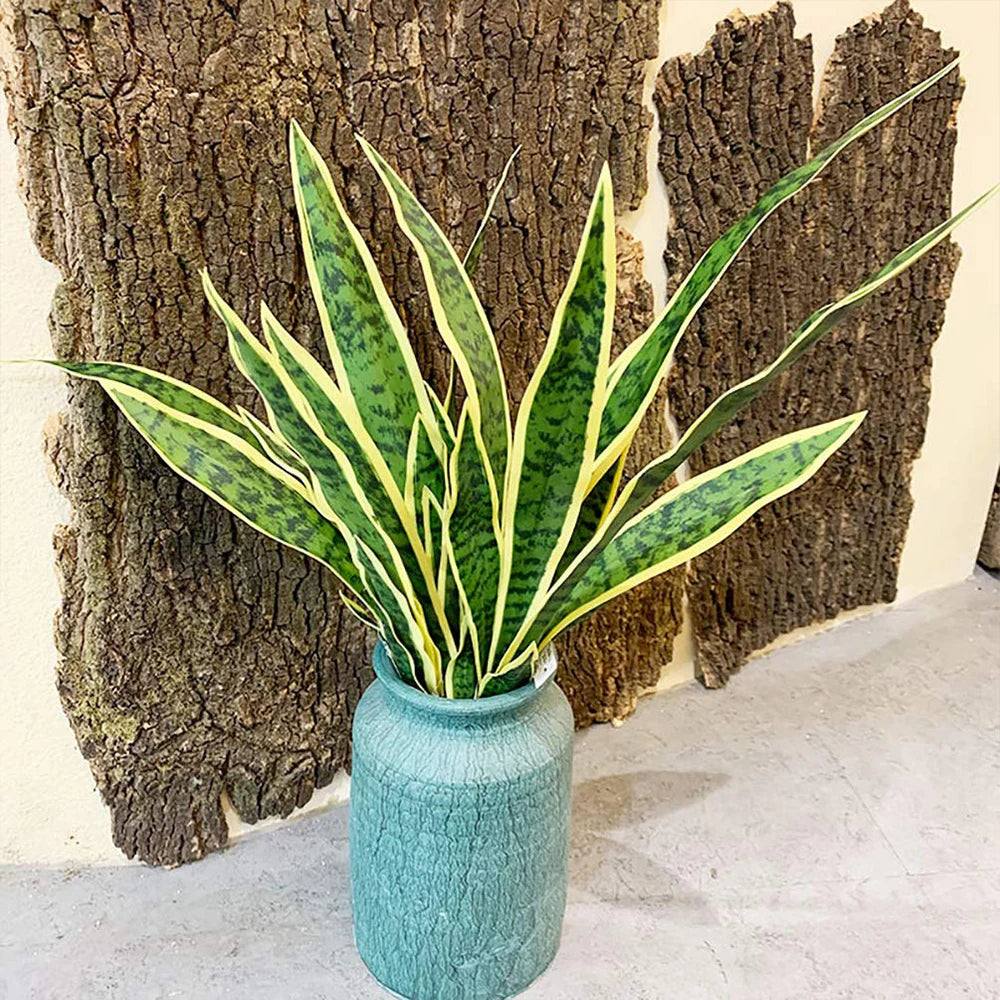 Una alta planta serpiente artificial en una maceta de cerámica verde azulada, exhibida contra un fondo de tronco de árbol tex