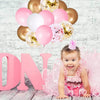 A baby girl is surrounded by pink and gold confetti latex balloons, with large pink letters spelling out 'DN' in the backgrou