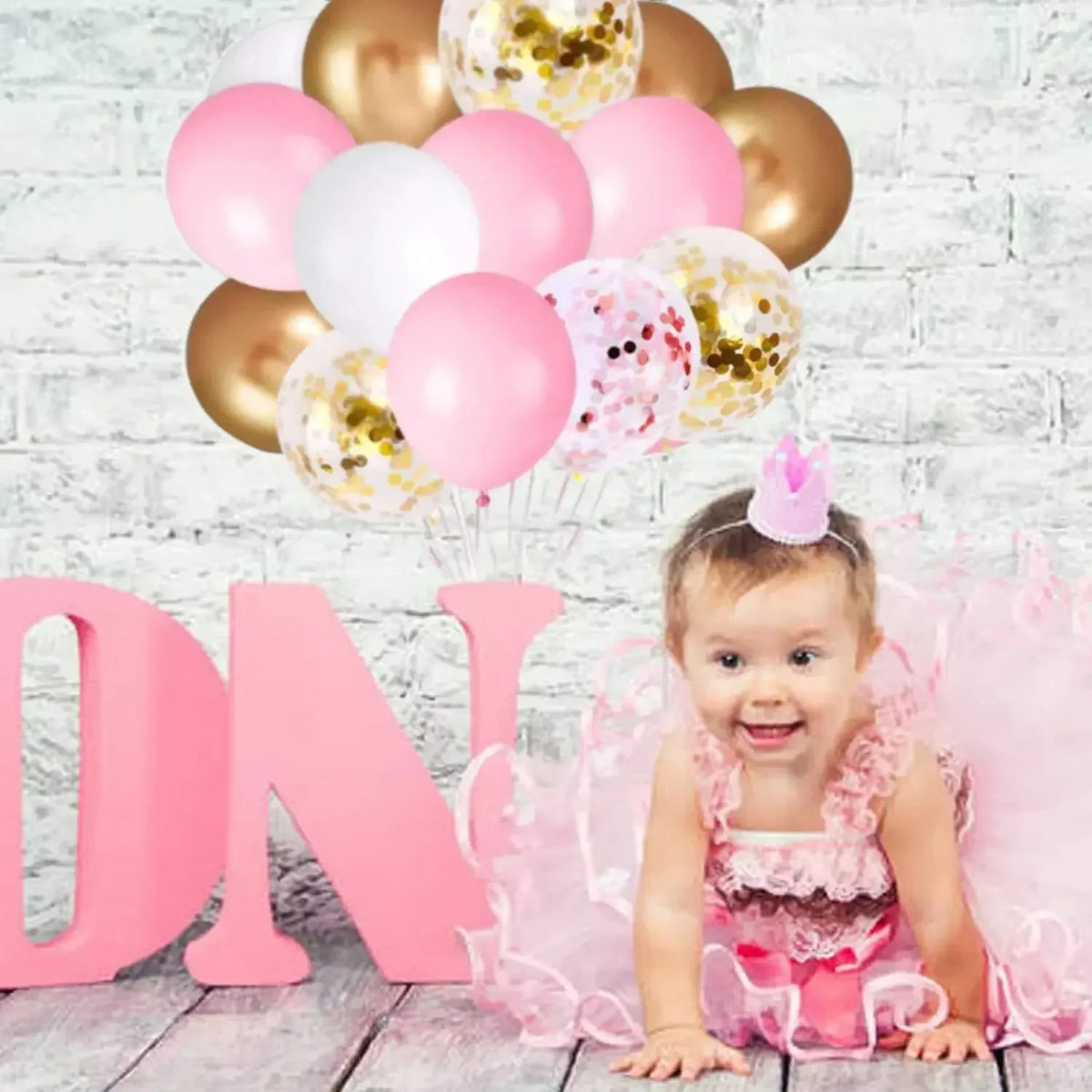 A baby girl is surrounded by pink and gold confetti latex balloons, with large pink letters spelling out 'DN' in the backgrou