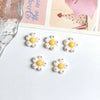 A set of six white and yellow flower resin nail art charms displayed on a white background next to a piece of paper.