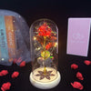 A rose crystal flower in an illuminated glass dome sits on a wooden base with scattered red rose petals.
