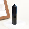 A black natural obsidian crystal obelisk point standing on a white surface.