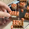 A hand places small, egg-shaped miniature food items into a wooden basket and a plastic tray.