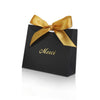 A black paperboard thank you gift bag tied with a gold ribbon, featuring the word Merci printed on it.