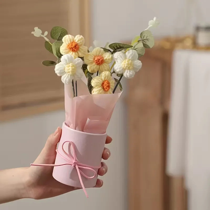 A hand holds a pink paper bouquet featuring yellow and white flowers with green leaves.