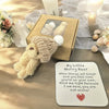 A cream-colored knitted bear plush toy rests on a wooden surface next to an encouraging card and a small gift box.