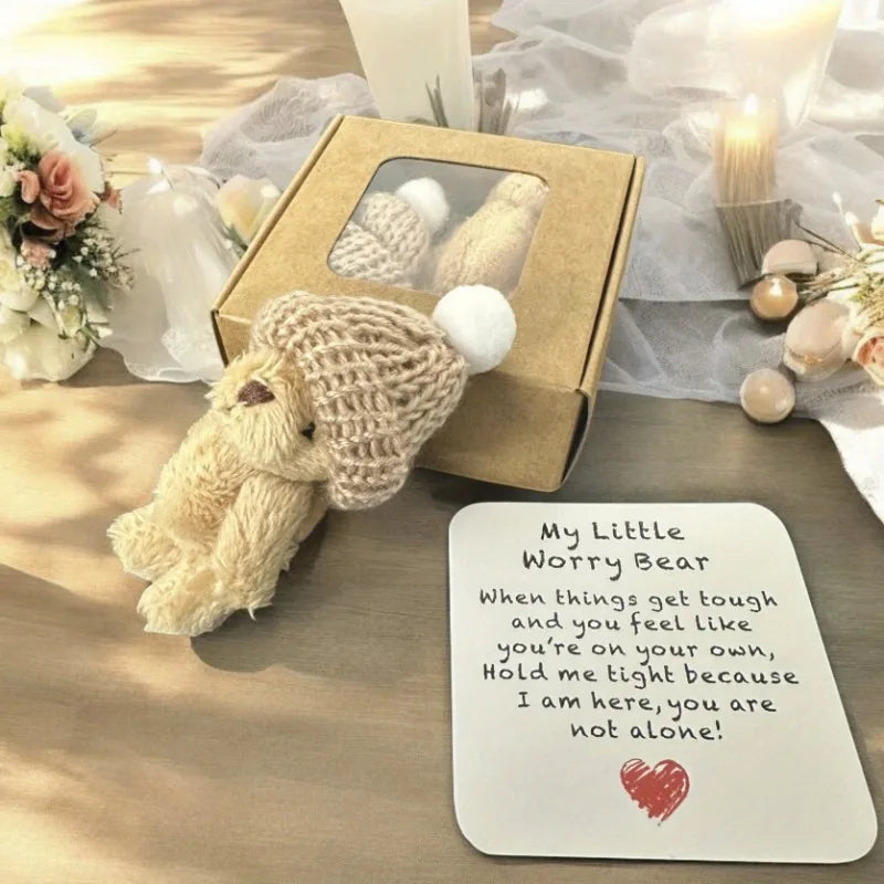 A cream-colored knitted bear plush toy rests on a wooden surface next to an encouraging card and a small gift box.