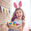 A young girl holding a woven basket filled with colorful Easter eggs and bunny ear headbands.