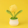 A hand-woven crochet flower in yellow sits in a white pot against a yellow background.