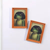 A framed illustration of a green frog in a wooden frame with a braided border.