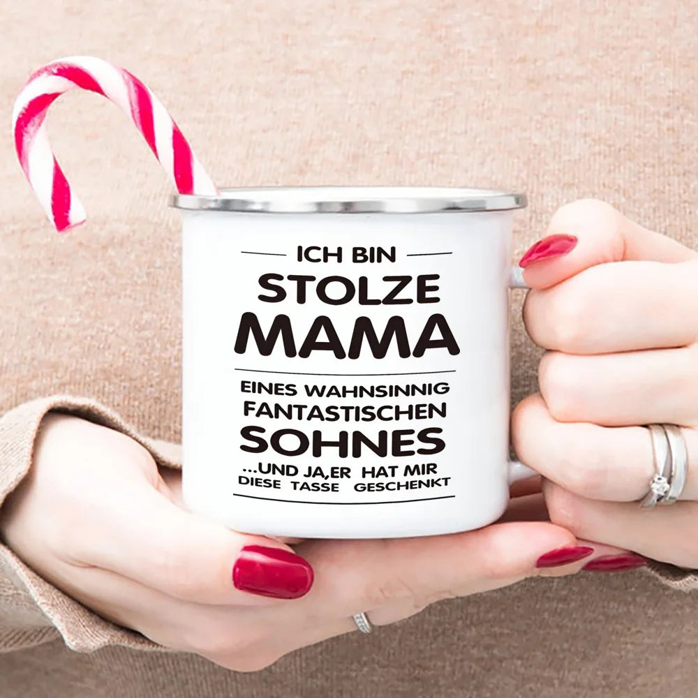A white enamel mug with black text reading Ich bin stolze Mama and a red and white striped candy cane is held by a person.