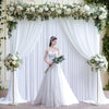 A bride stands in a white wedding dress in front of a sheer white fabric backdrop decorated with greenery and roses.