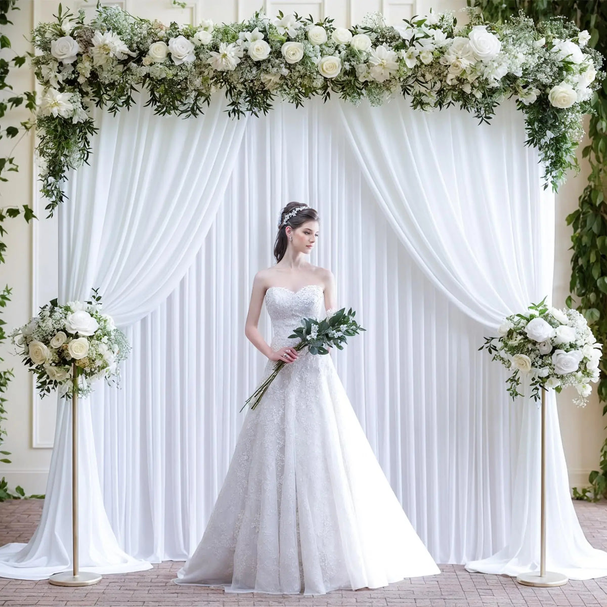 A bride stands in a white gown against a draped satin backdrop decorated with white flowers and greenery.