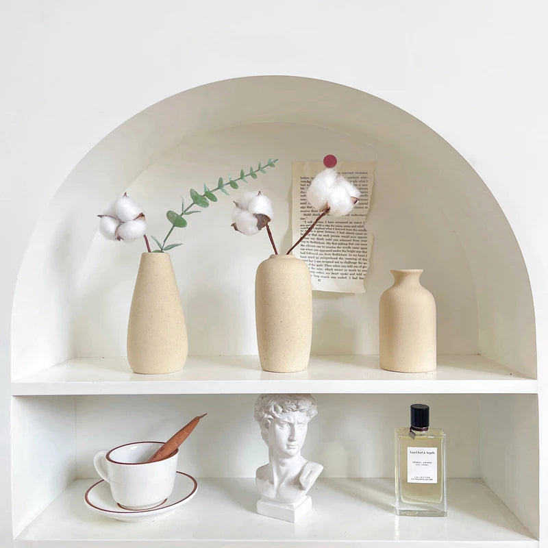 A minimalist ceramic vase is displayed on a white shelf within an arched niche, surrounded by dried greenery and white flower