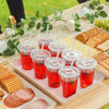 A tray of red fruit drinks served in clear disposable plastic cups with flat lids, surrounded by crackers and grapes.