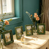 A vintage Baroque resin picture frame with a green and gold frame sits on a light table, surrounded by roses and greenery.