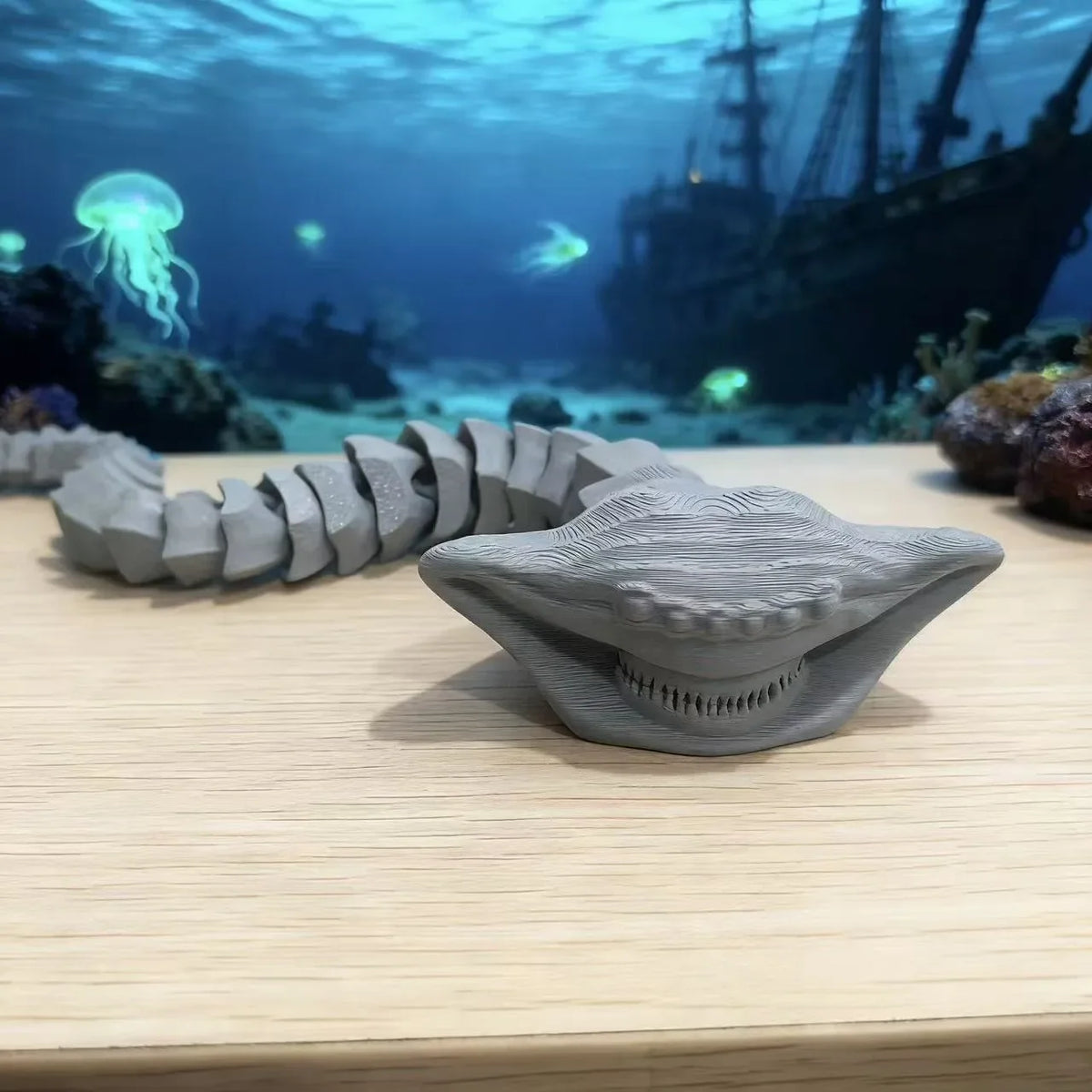 A gray 3D printed articulated sea monster fidget toy resting on a wooden surface with an underwater background.