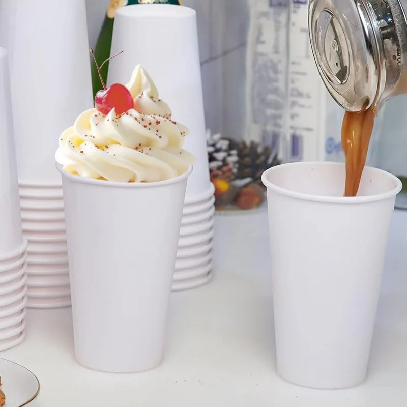 A white disposable paper coffee cup filled with whipped cream and a cherry, next to a metal coffee dripper.