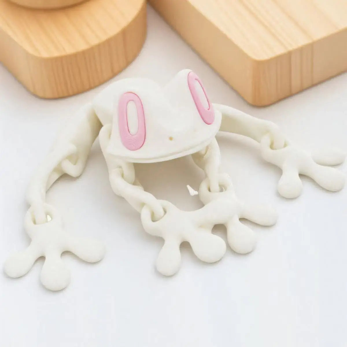 A white and pink tree frog ornament with long limbs is displayed on a white surface next to wooden blocks.