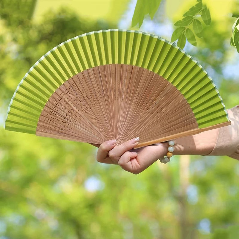 A hand holding an elegant bamboo folding fan with green slats and a tassel against a bright, leafy green background.