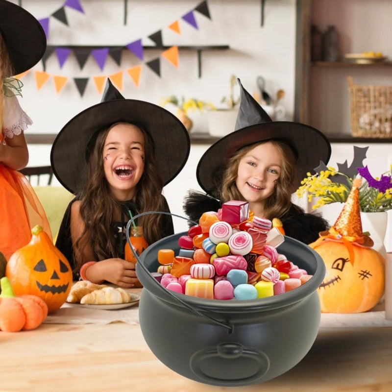 A black witch cauldron bowl filled with colorful candies sits on a wooden table next to a child wearing a witch hat.