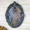 A dark, ornate mirror frame shaped like a face surrounded by vines and leaves, featuring a hanging pentagram amulet.