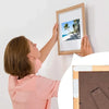 A woman is hanging a framed picture using reusable double-sided adhesive strips on a white wall.