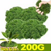 A hand holds a clump of dark green artificial moss, with a potted plant visible below.
