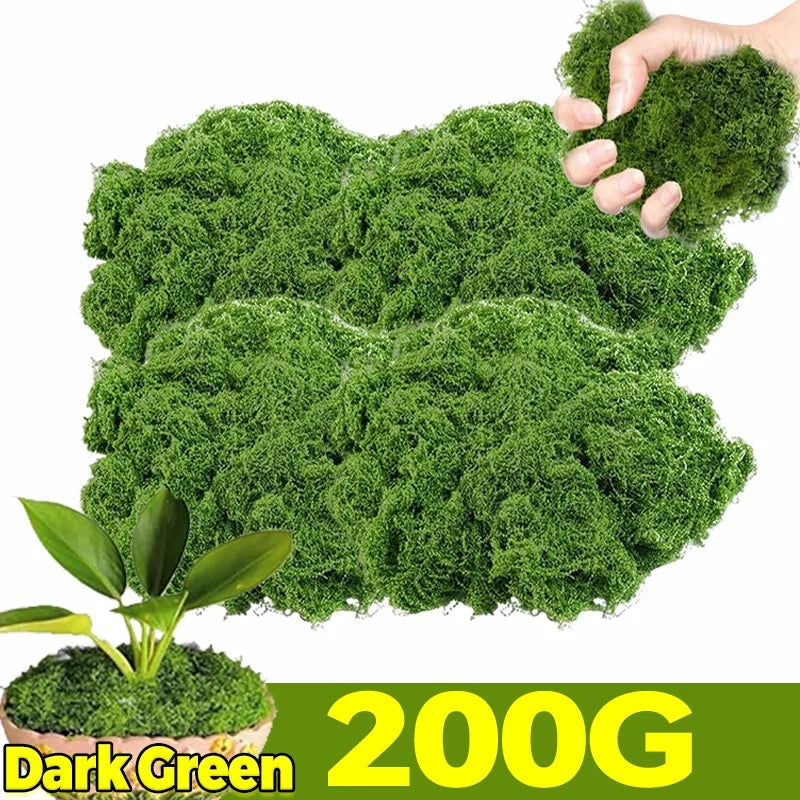 A hand holds a clump of dark green artificial moss, with a potted plant visible below.