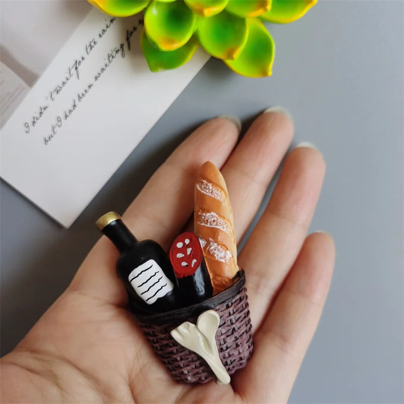 A hand holds a small basket of food-shaped fridge magnets including a baguette and a tomato.