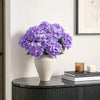A bouquet of purple artificial hydrangea silk flowers in a white vase on a dark marble surface.