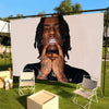 A man with dreadlocks is pictured against a white backdrop, making a hand gesture, seated outdoors on green grass.