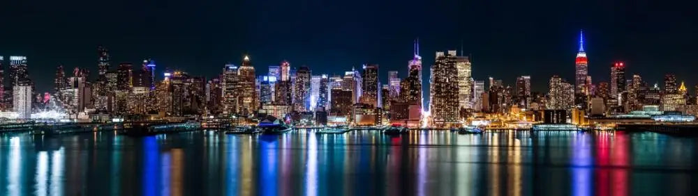 A nighttime view of the New York City skyline reflected on water.