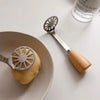 A silver manual masher with a wooden handle is shown next to a piece of potato on a ceramic plate.