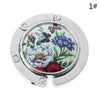 Silver foldable handbag hook with a floral design for securing bags on tables.