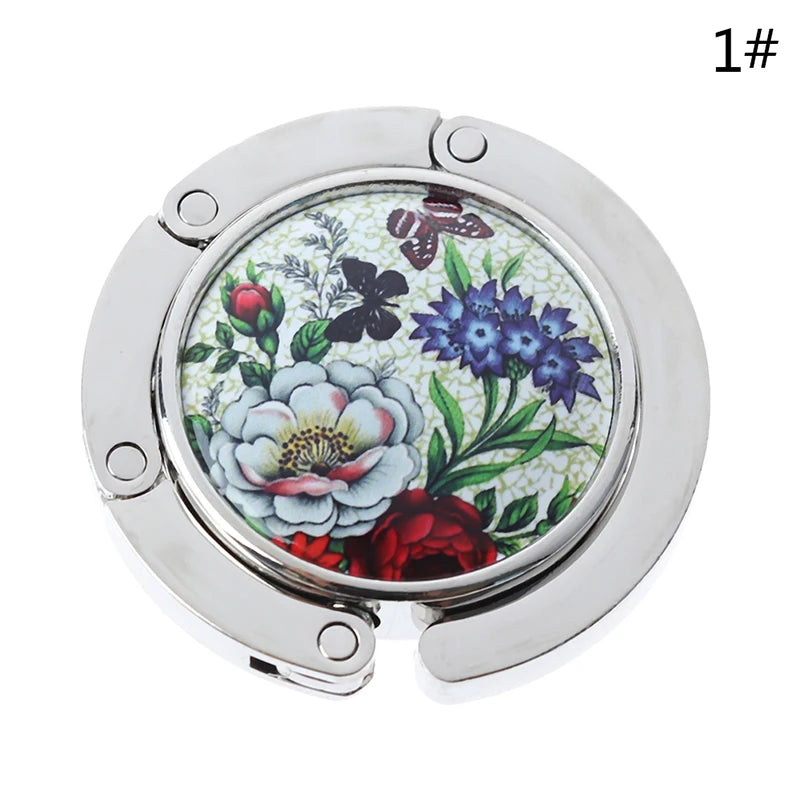 Silver foldable handbag hook with a floral design for securing bags on tables.